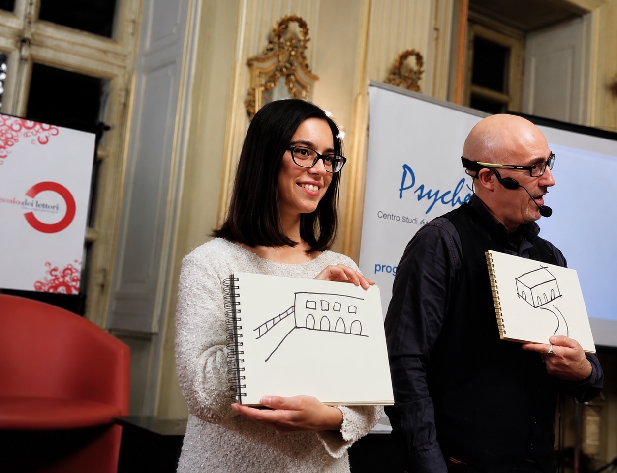 Antonio Argus con microfono ad archetto durante un intervento pubblico in una sala conferenze, con un partecipante e un disegno su blocco note